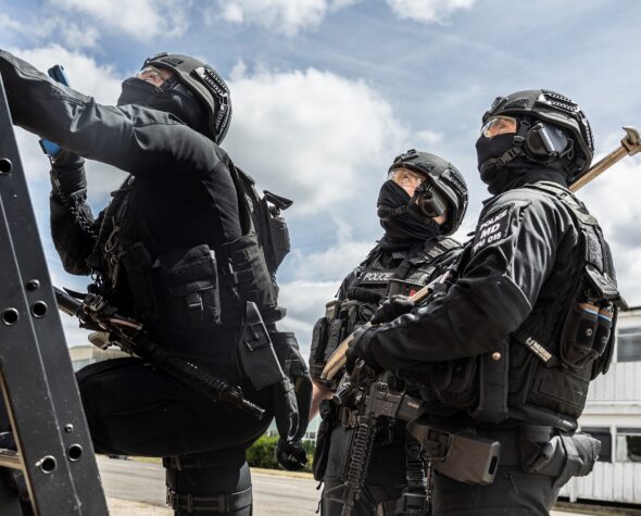 Two Ministry of Defence Police Tactical Firearms Unit officers standing in front of a ladder whilst a colleague climbs the ladder